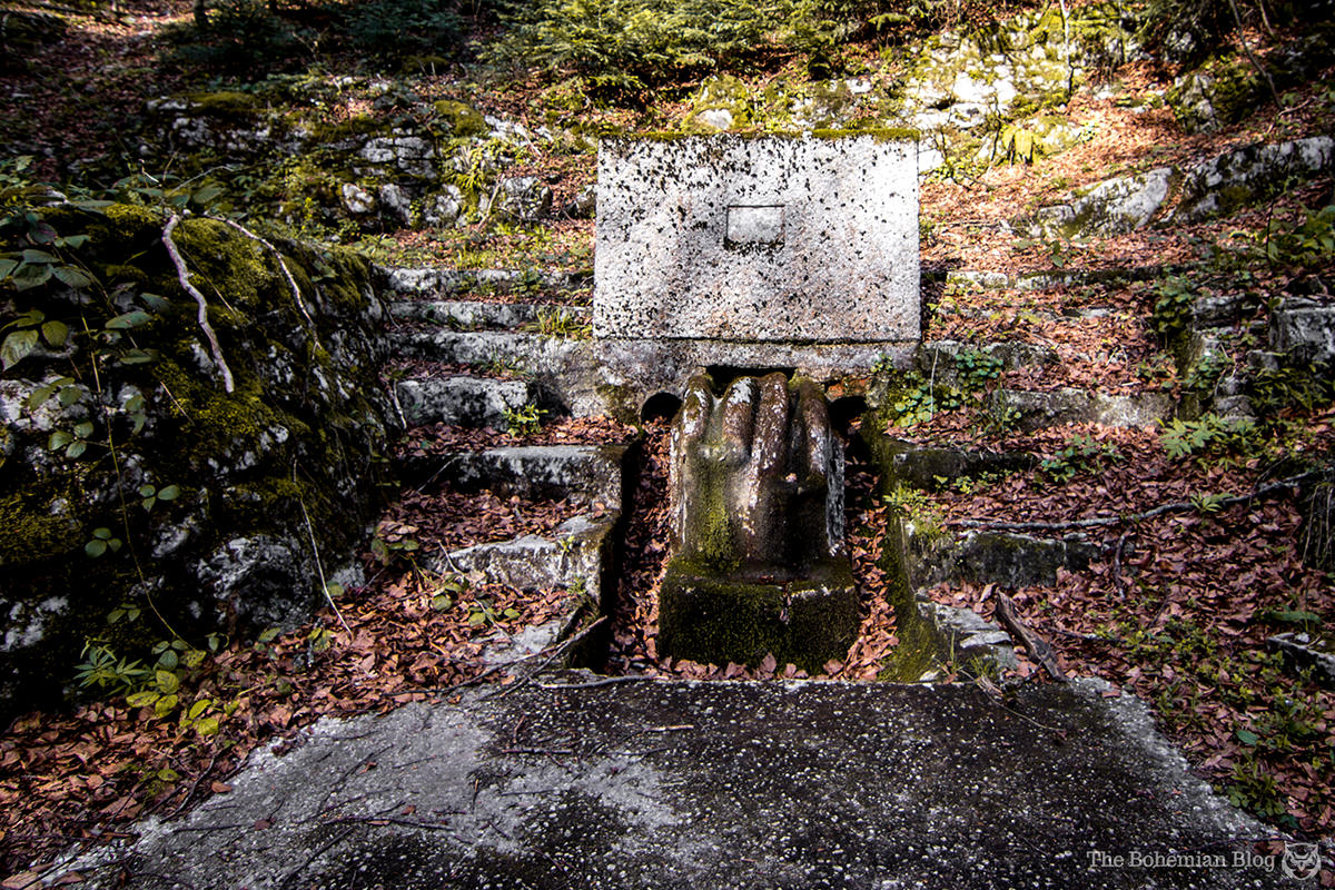 A strange stone fountain lies covered in moss and leaves, in a meadow behind the monument.